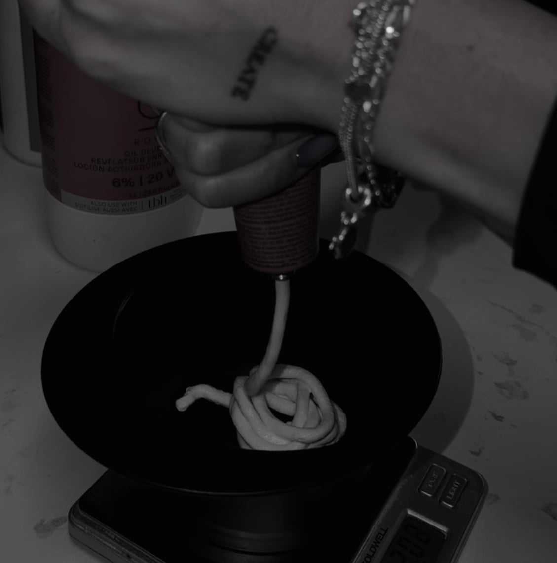 Mixing hair dye in a black bowl on a digital scale, close-up of hand with tattoos and bracelets.