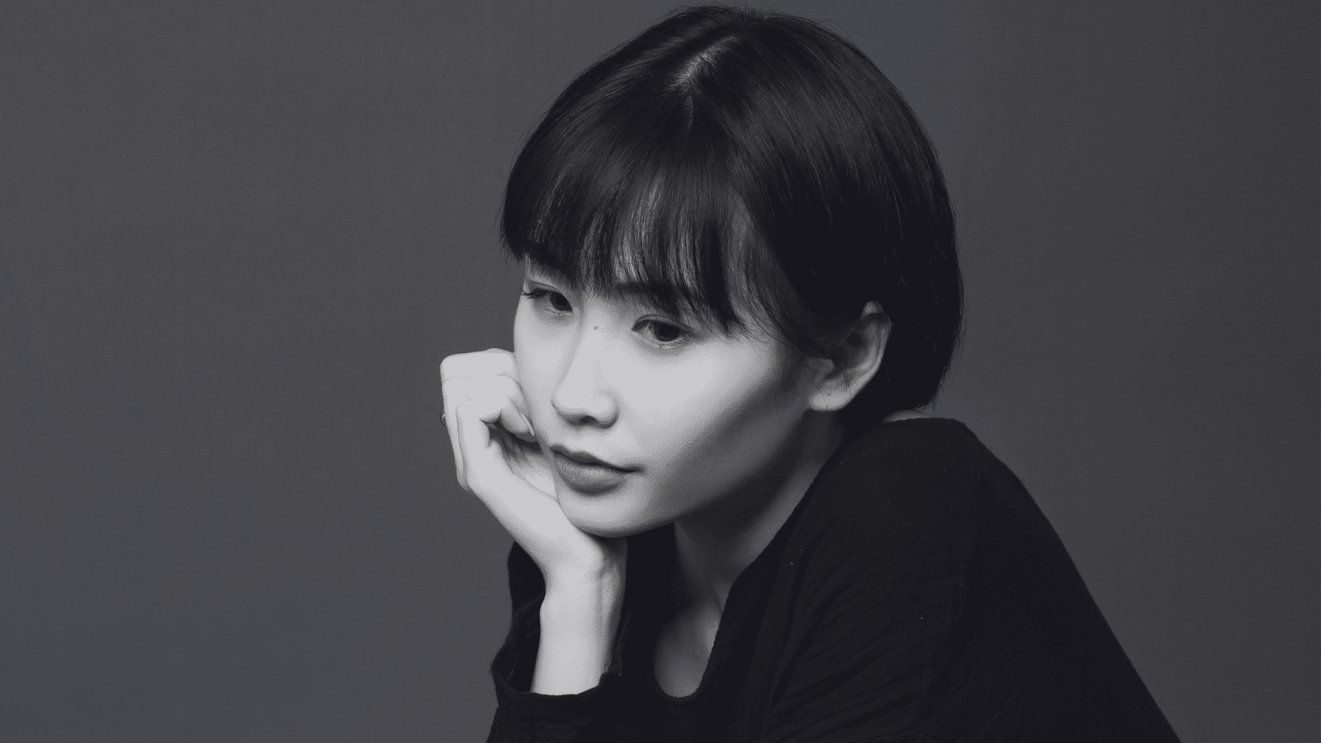 A contemplative person with short hair, resting their chin on hand, in a grayscale portrait.