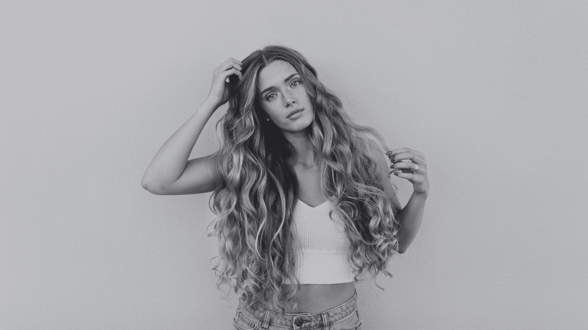 Youthful woman with long wavy hair, wearing a white tank top and jeans against a plain backdrop.